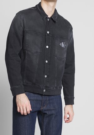 Mężczyzna noszący czarną kurtkę jeansową zapinaną na guziki z kieszeniami na piersi i logo Calvin Klein, w połączeniu z ciemnoniebieskimi dżinsami.