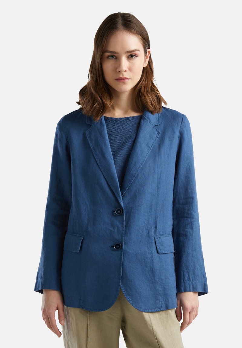 Blazer azul de lino con un diseño ajustado, dos botones, solapas con muesca y dos bolsillos frontales. Lleva una blusa azul texturizada debajo.