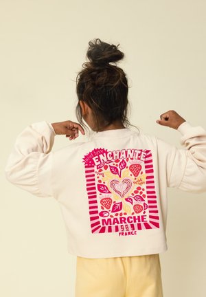 Kind met haar in een knot, draagend een crèmekleurige trui met een roze en rode afbeelding van aardbeien, bladeren en de woorden "Enchanté Marché France."