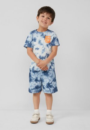 Niebieska koszulka tie-dye z pomarańczowym napisem, w zestawie z dopasowanymi szortami tie-dye. Strój ma krótkie rękawy i luźny krój.