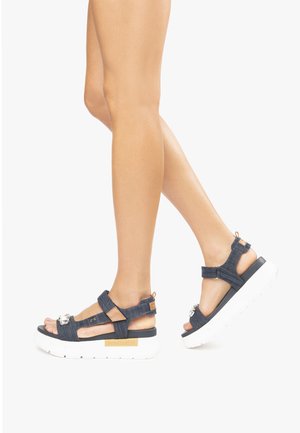 Denim platform sandals with adjustable straps, a white thick sole, and decorative hardware. Features a dark blue color and a casual design.