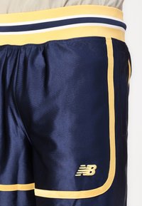 Shorts en polyester bleu marine avec une ceinture élastique rayée jaune et bleue, présentant une bordure jaune et un logo New Balance en bas à gauche.