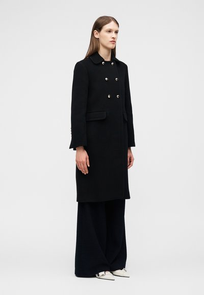 Frau, die einen langen schwarzen zweireihigen Mantel mit silbernen Knöpfen, schwarze weit geschnittene Hosen und weiße spitze Schuhe trägt, vor einem schlichten Hintergrund stehend.
