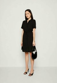 Črna obleka s kratkimi rokavi, pasom za zavezovanje in razširjenim robom. V kombinaciji s črnimi petkami in sijajno torbico, ki prikazuje eleganten dizajn.