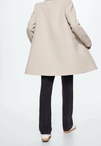 Beige uldfrakke med en flagrende design, rygslids og to sidelommer, parret med mørke jeans og lyse sko.