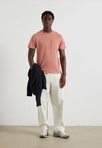 Lichtroze t-shirt met korte mouwen en een borstzak, gecombineerd met losse witte broek en grijze sneakers. Een marineblauw sweater hangt over één arm.