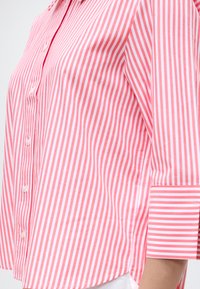 Ροζ και λευκό πουκάμισο με κάθετες ρίγες, κουμπωτό με τρία τέταρτα μανίκια και αντίθετες οριζόντιες ρίγες στις μανσέτες.