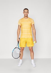 Gul atletisk T-skjorte med horisontale striper og Nike-logo, kombinert med matchende gule shorts, holder en tennisracket og ball.