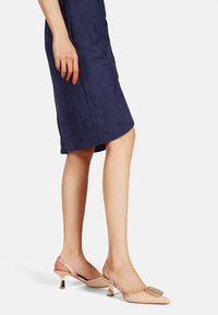 Décolleté beige con cinturino alla caviglia, punta appuntita, trama in rete, grande fibbia decorativa e tacco metallico. Indossato con un vestito blu navy.