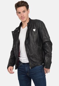 Zwarte leren jas met ritssluiting, gequilt schouderdetails en logo-accent. Draagt over een wit shirt, gecombineerd met blauwe jeans.