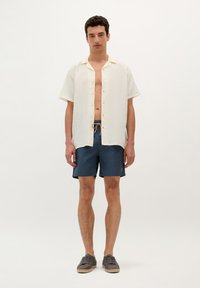 Homme portant une chemise crème à manches courtes ouverte, un short bleu marine à cordon de serrage et des espadrilles grises, se tenant devant un fond blanc uni.