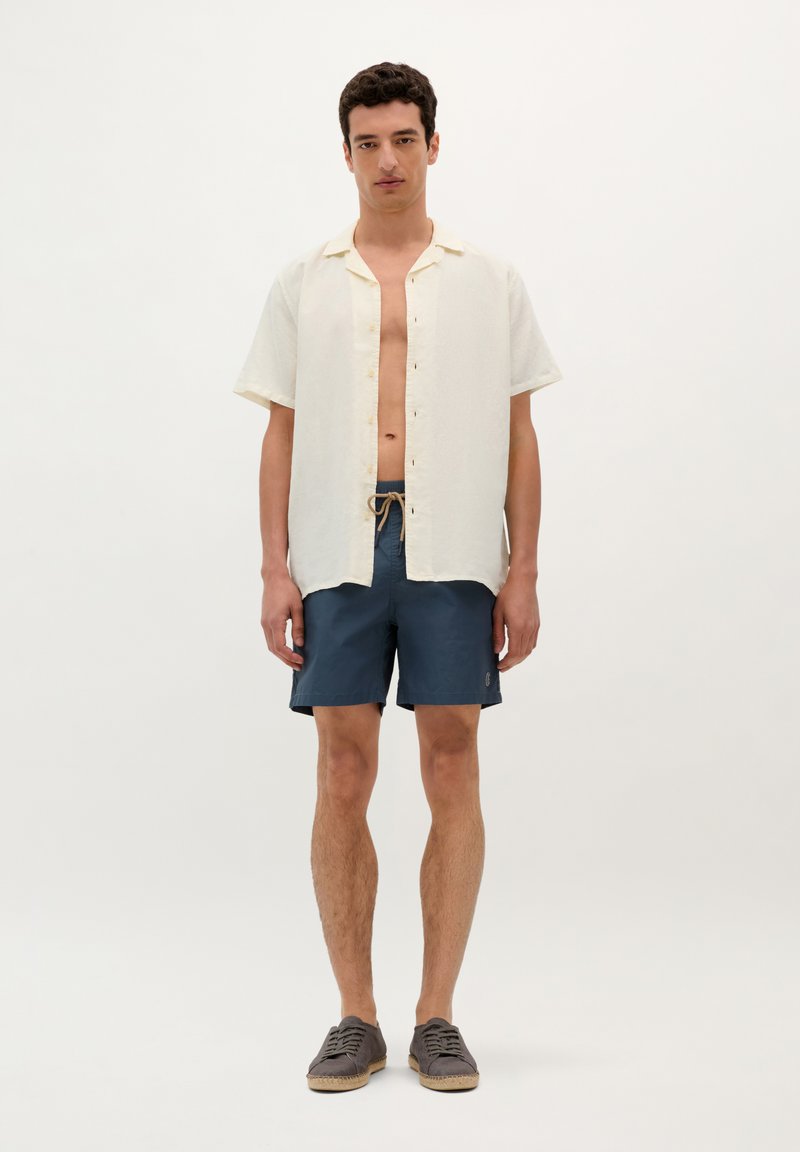 Homme portant une chemise crème à manches courtes ouverte, un short bleu marine à cordon de serrage et des espadrilles grises, se tenant devant un fond blanc uni.