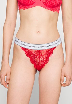 Red lace thong featuring a floral design, with a white waistband embossed with "GUESS." The fabric has a delicate texture and intricate detailing.