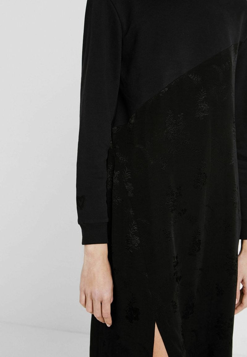 Zwarte jurk met een gestructureerde, bloemige onderpaneel en een solide bovenstuk. Lange mouwen met geribbelde manchetten. Zijslitdetail.