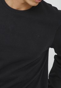 Felpa nera in cotone a manica lunga con scollo a girocollo a coste. Presenta un piccolo logo discreto sul petto. Tessuto morbido, maniche di vestibilità standard.