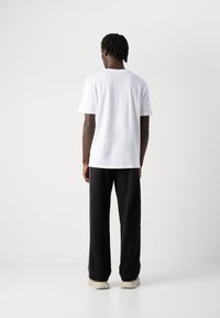 Mann som står med ryggen til, iført en enkel hvit t-skjorte, svarte vide bukser og lyse sneakers mot en enkel hvit bakgrunn.