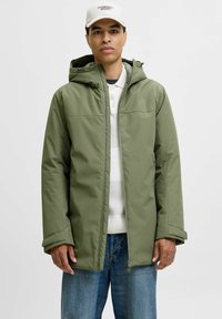 Olivgrüne wasserdichte Jacke mit Kapuze, Reißverschluss und Seitentaschen, getragen über einem weißen Pullover, kombiniert mit blauer Jeans.