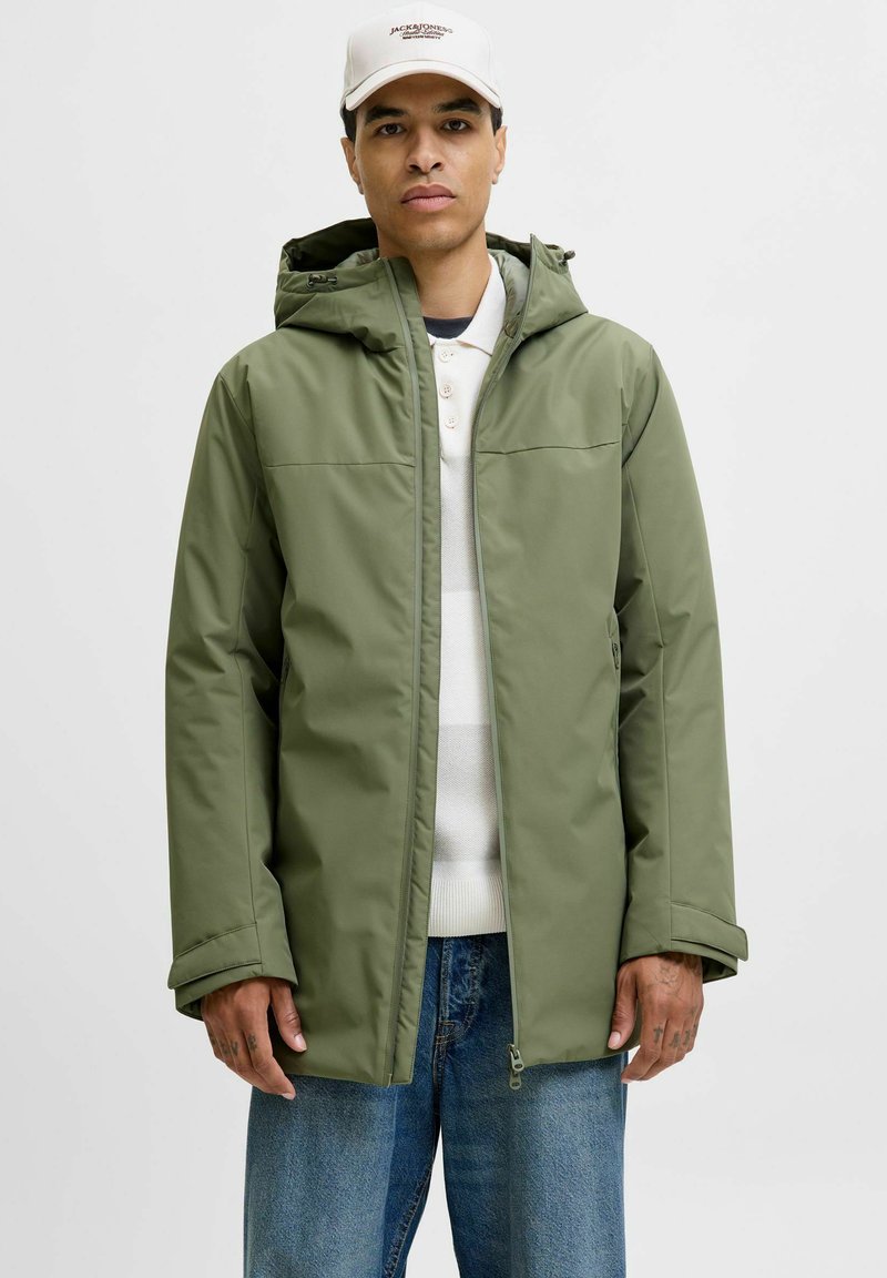 Olivgrüne wasserdichte Jacke mit Kapuze, Reißverschluss und Seitentaschen, getragen über einem weißen Pullover, kombiniert mit blauer Jeans.