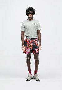 Γκρι κοντομάνικη μπλούζα Nike, πολύχρωμοι σορτς με αφηρημένα σχέδια, κόκκινες κάλτσες, γκρι αθλητικά παπούτσια και σκούρα γυαλιά ηλίου, στέκεται μπροστά σε απλό φόντο.