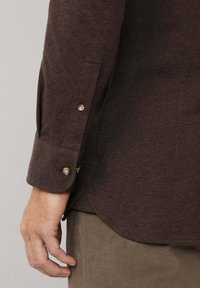 Acercamiento de un brazo izquierdo de una persona que lleva una camisa de manga larga marrón oscuro con puño abotonado y pantalones caqui.