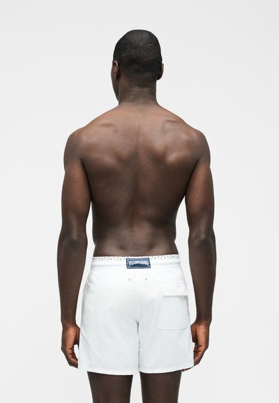 Άνδρας με σκουρόχρωμο δέρμα, ο οποίος φαίνεται από πίσω, φορώντας λευκά μαγιό σορτς με πίσω τσέπη και ζώνη μέσης με λογότυπο.