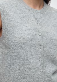 Gros plan d'un gilet en maille duveteuse gris clair sans manches avec sept petits boutons ronds sur le devant.