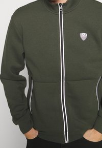 Mörkgrön zip-up-tröja med hög krage, vit dragkedja och logopatch. Har horisontell söm och sidostråk.