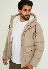 Beige gepolsterte Jacke mit abnehmbarer Kapuze, weißem Kunstfellfutter, frontalen Taschen und schwarzen Kordeln, kombiniert mit einem weißen T-Shirt.