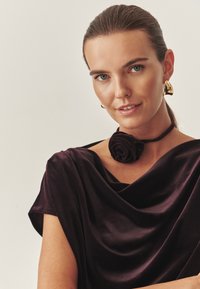 Sort fløjlskjole med en draperet halsudskæring, prydet med en krave lavet af et stofrose. Guld geometriske øreringe tilføjer kontrast.