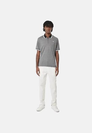 Joven con cabello oscuro que lleva una camisa polo gris, pantalones blancos y zapatillas blancas, de pie frente a un fondo claro y sencillo.