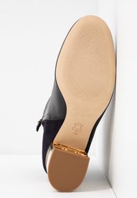 Semelle d'une botte cheville en cuir noir avec semelle en cuir beige texturée, talon bloc avec accents dorés et fermeture éclair latérale visible. Taille indiquée 8M.