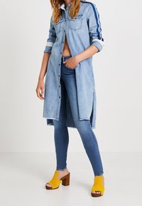 Vestido camisero de denim con dos bolsillos frontales, rayas azules en las mangas, aberturas laterales, combinado con jeans ajustados azules y sandalias de tacón amarillas.