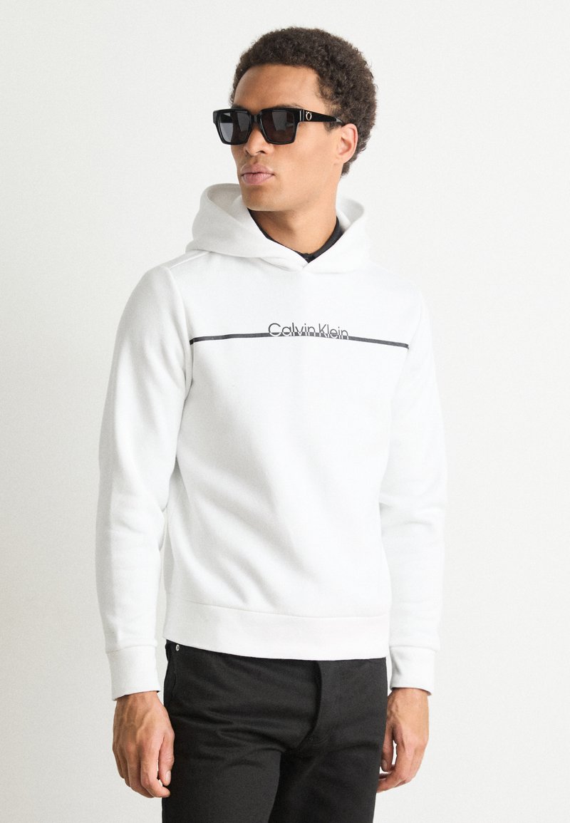 Hanorac alb cu glugă, decorat cu o dungi orizontale negre și logo-ul "Calvin Klein", asortat cu pantaloni închiși la culoare și ochelari de soare negri.