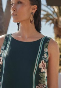 Ärmelloses Kleid mit Blumenmuster in dunkelgrün und mehrfarbigen Streifen. Das Model trägt große, geschmückte Ohrringe mit roten Akzenten.