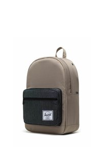 Sac à dos en tissu beige de forme arrondie, avec une poche avant zippée noire et un patch logo blanc. Bretelles réglables et poignée sur le dessus.