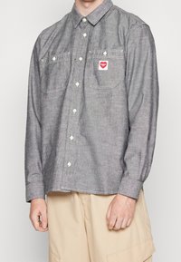 Man draagt een grijs overhemd met knopen, borstzakken en een klein rood hartvormig patch, gecombineerd met beige cargoshorts.