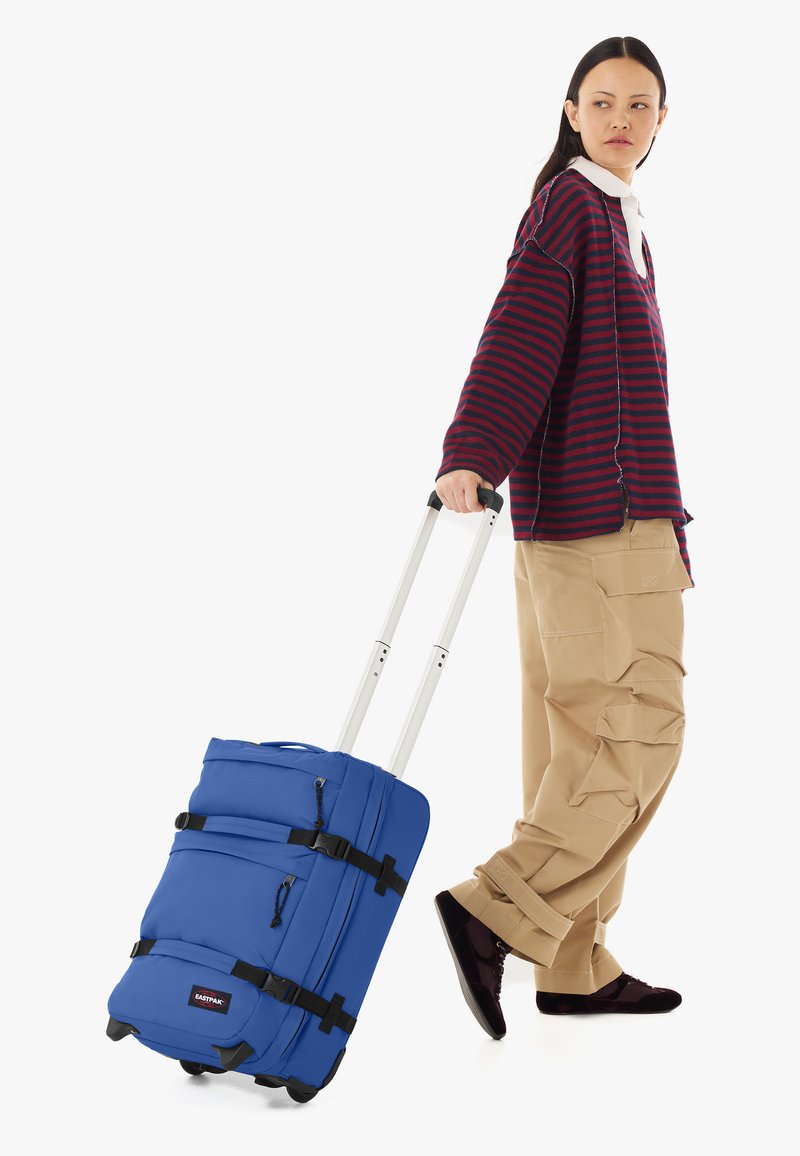 Personne portant une chemise à rayures à manches longues et un pantalon cargo beige tire une grande valise bleue à roulettes avec une poignée extensible.