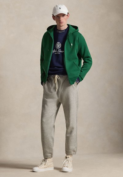 Mees, kellel on peas valge nokamüts, roheline tõmblukuga kapuutsiga pusa, tumesinine dressipluus, hallid dressipüksid ja beežid kõrge säärega tossud, seisab ühevärvilise tausta ees.