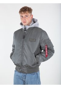 Giacca bomber grigia con una finitura lucida, polsini a coste e zip frontale. Include un cappuccio grigio e un'etichetta di tessuto rossa sulla manica.