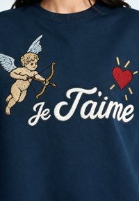 Un chérubin avec un arc et une flèche visant un cœur rouge à côté de la phrase brodée « Je T’aime » sur un tissu bleu marine.
