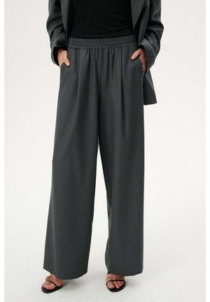 Osoba ubrana w ciemnoszare szerokie spodnie z elastycznym pasem, czarną marynarkę, czarny top i czarne sandały z odkrytymi palcami. Ręce w kieszeniach.