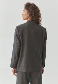 Charcoalgrijze op maat gemaakte blazer van een gestructureerde stof, met klassieke revers en knoopmanchetten. Zicht vanaf de achterkant, die een rechte snit toont.