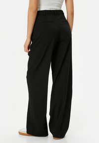 Zwarte wijde broek gemaakt van gladde stof, met een hoge taille, twee achterzakken en een relaxte pasvorm. Paar witte sneakers zichtbaar.