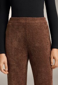 Osoba nosi smeđe hlače visokog struka s teksturom i crnu majicu dugih rukava, prikazuje detalje od struka do sredine bedra.