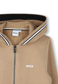 Haut à capuche zippé marron avec un col côtelé noir et blanc. Il dispose d'une fermeture éclair à l'avant et d'un logo "BOSS" blanc sur la poitrine.