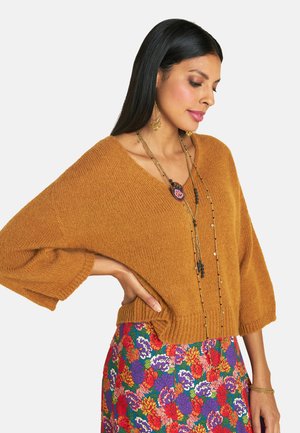 Senfgelber gestrickter Pullover mit weiten Ärmeln und V-Ausschnitt, kombiniert mit einem bunten Rock mit Blumendruck. Eine lange mehrlagige Halskette betont den Look.
