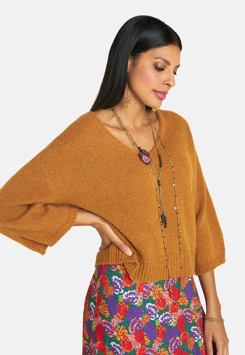 Senfgelber gestrickter Pullover mit weiten Ärmeln und V-Ausschnitt, kombiniert mit einem bunten Rock mit Blumendruck. Eine lange mehrlagige Halskette betont den Look.