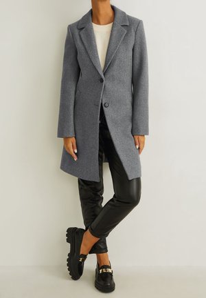 Femme portant un manteau en laine gris boutonné sur un top blanc, un pantalon en cuir noir et des mocassins noirs avec des semelles épaisses et des boucles dorées.