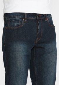 Jeans de mezclilla oscura con un ligero degradado, que presentan costuras de color cobre, cinco bolsillos y un cierre de botón en la cintura.
