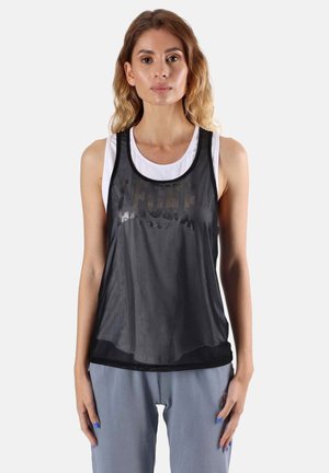 Femme aux cheveux ondulés mi-longs, portant un débardeur blanc sous un haut sans manches en maille noire et un pantalon bleu clair, face à l'avant.